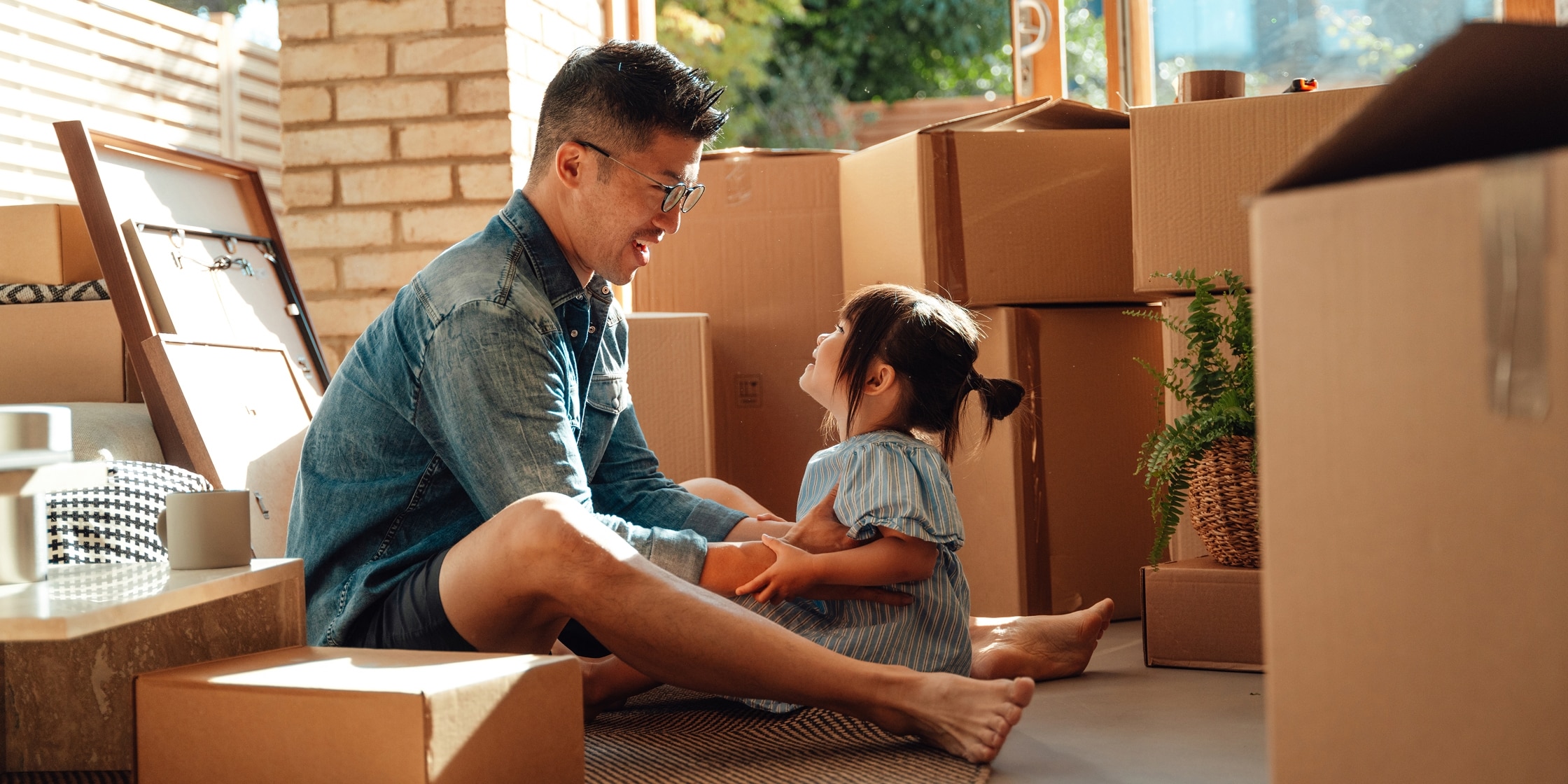 A father and his young daughter sit on the floor smiling at one another, surrounded by moving boxes.