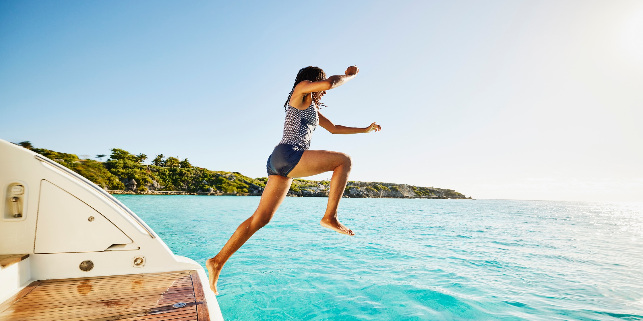 A woman leaps off the back of a boat into bright blue water set against a tropical rocky coastline.