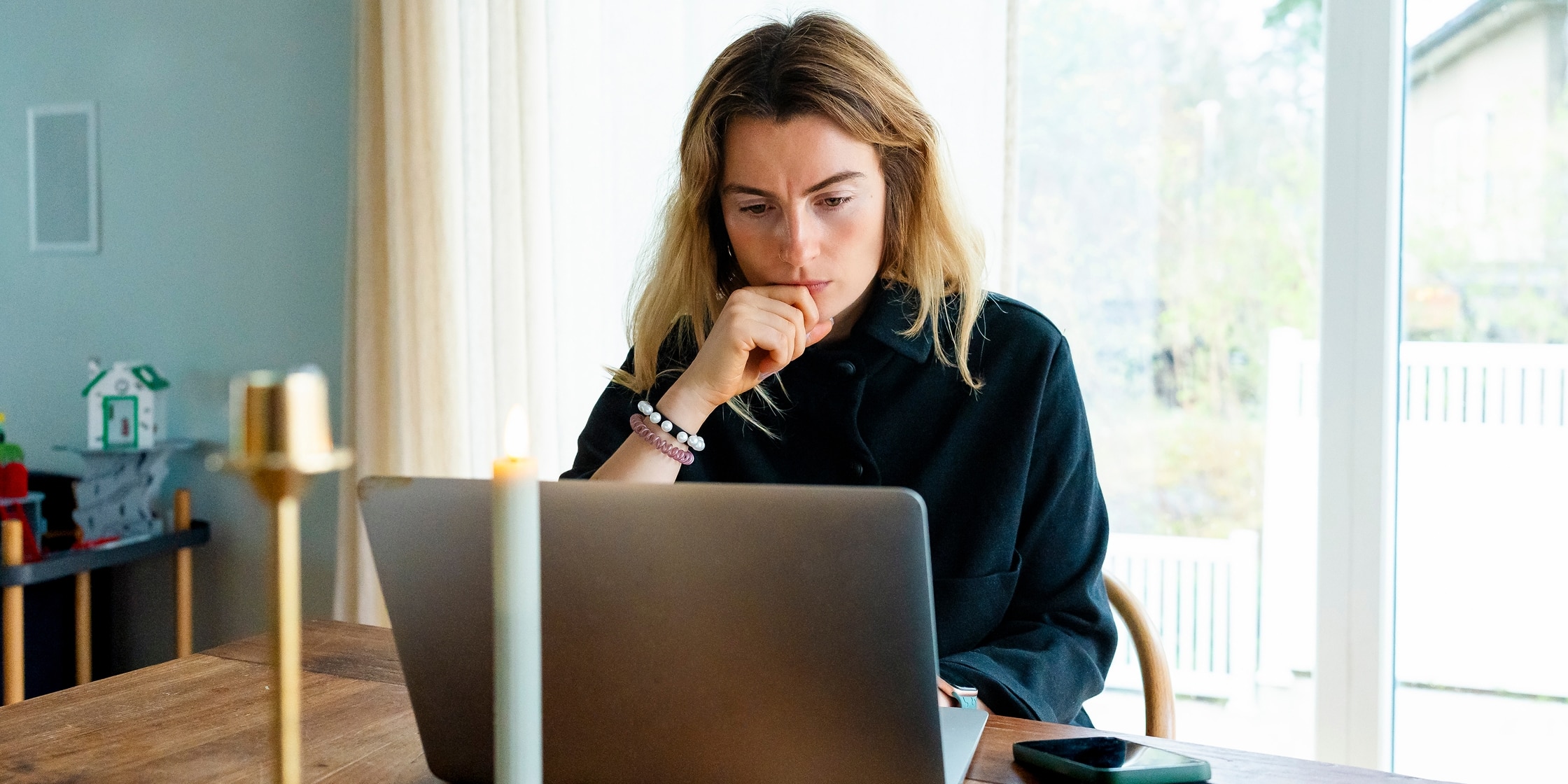 A woman lost in thought stares intently at her laptop while seated at her kitchen table.