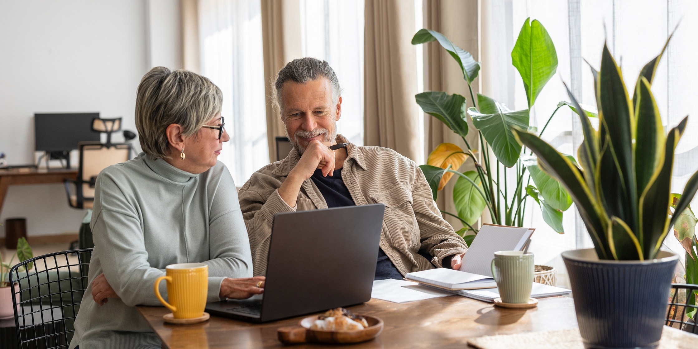 A senior couple sits at their kitchen table and reviews finances on their laptop.