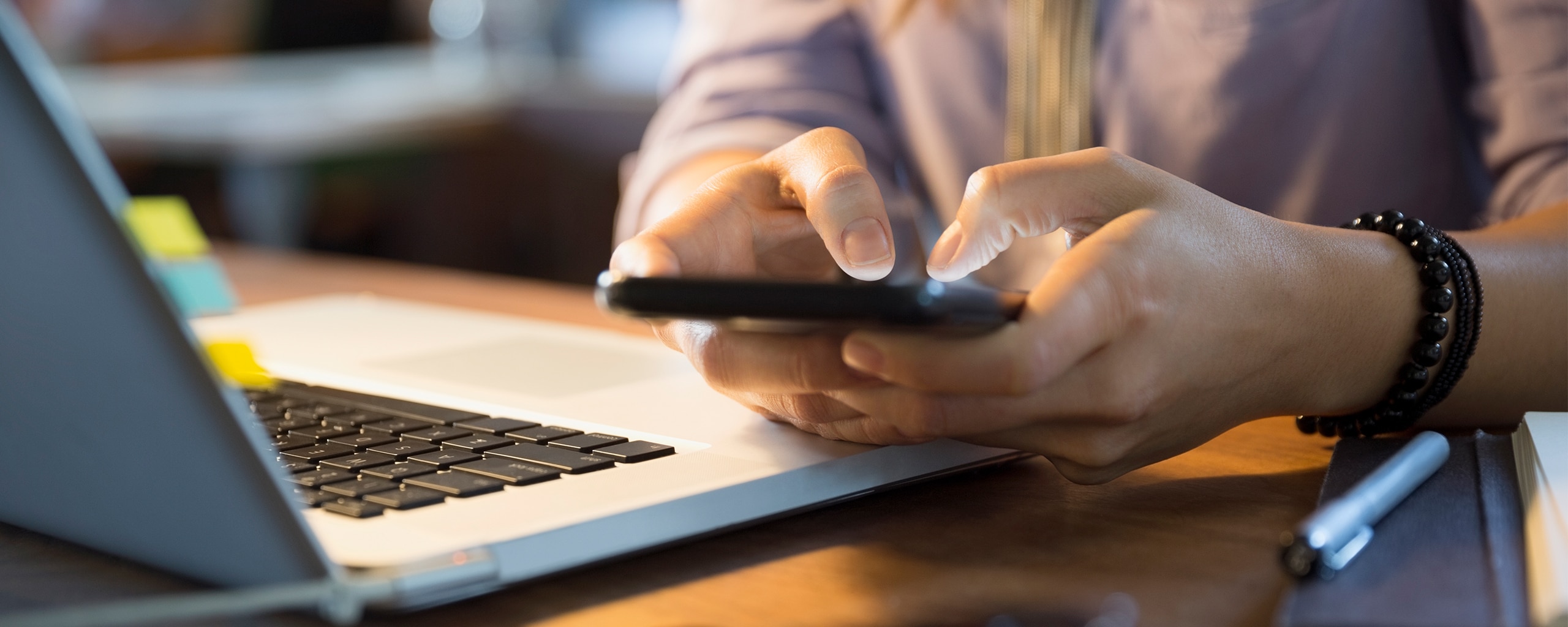 Close-up of hands using a smartphone beside a laptop.