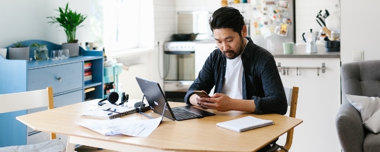 Someone sitting at their kitchen table while using a laptop and holding a phone.