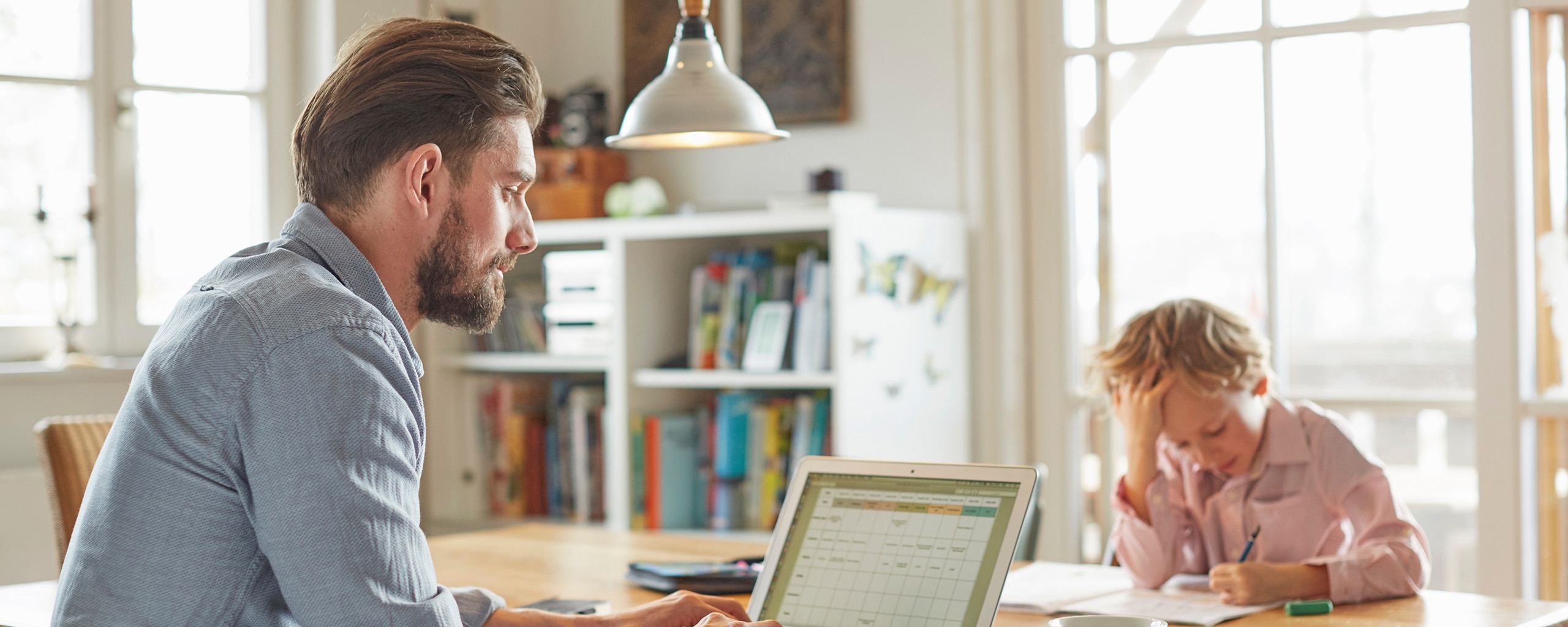 A man on a laptop works on a budget while a child does homework across the table.