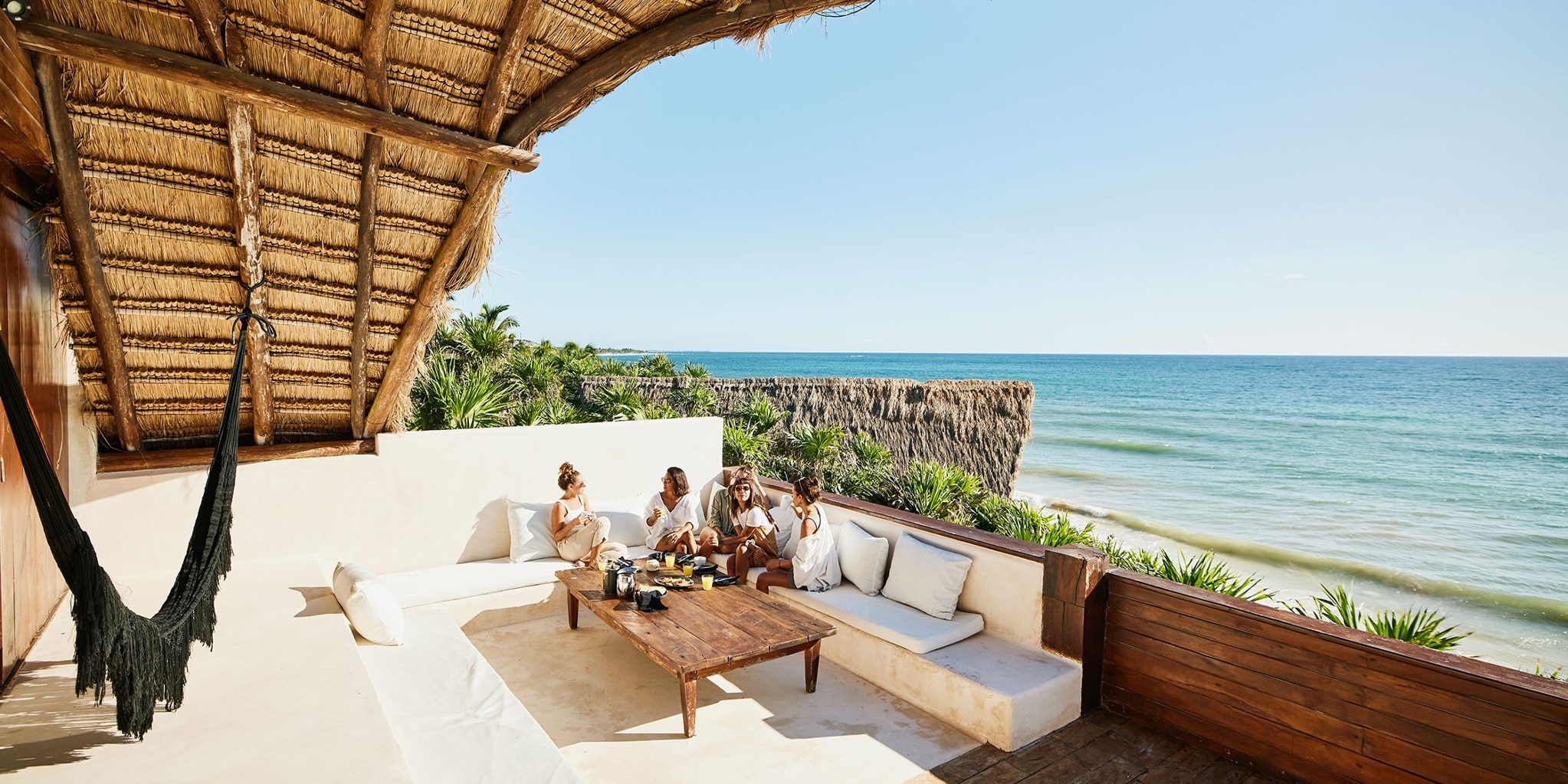 A group of vacationers sits in an outdoor lounge area that looks out over trees and an ocean.