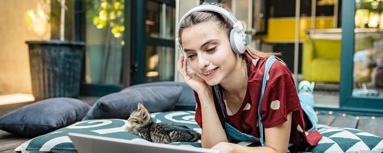 A person laying down next to a cat looks at an open screen with headphones on.