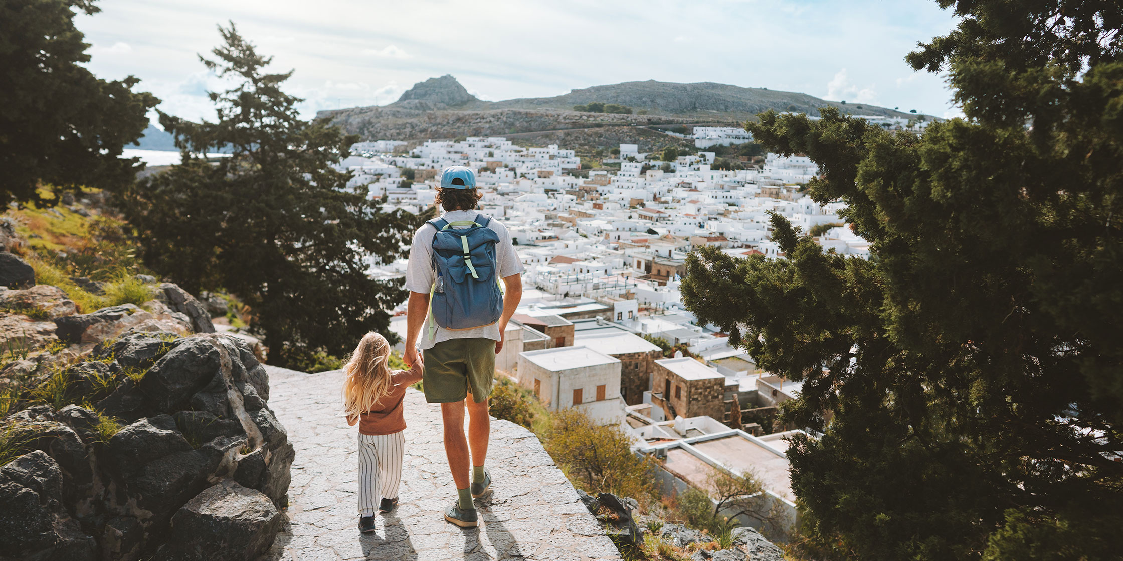 A man and child hold hands as they walk along a path high above a Greek town.