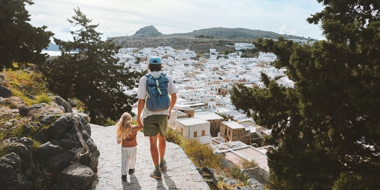 A person and child hold hands as they walk along a path high above a Greek town.