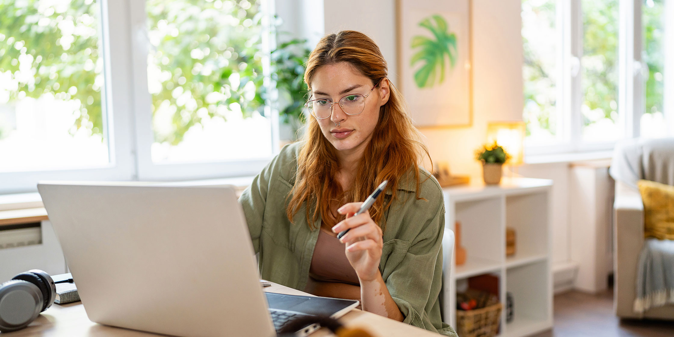 Young woman holds a pen as she sits at a table looking at her laptop.