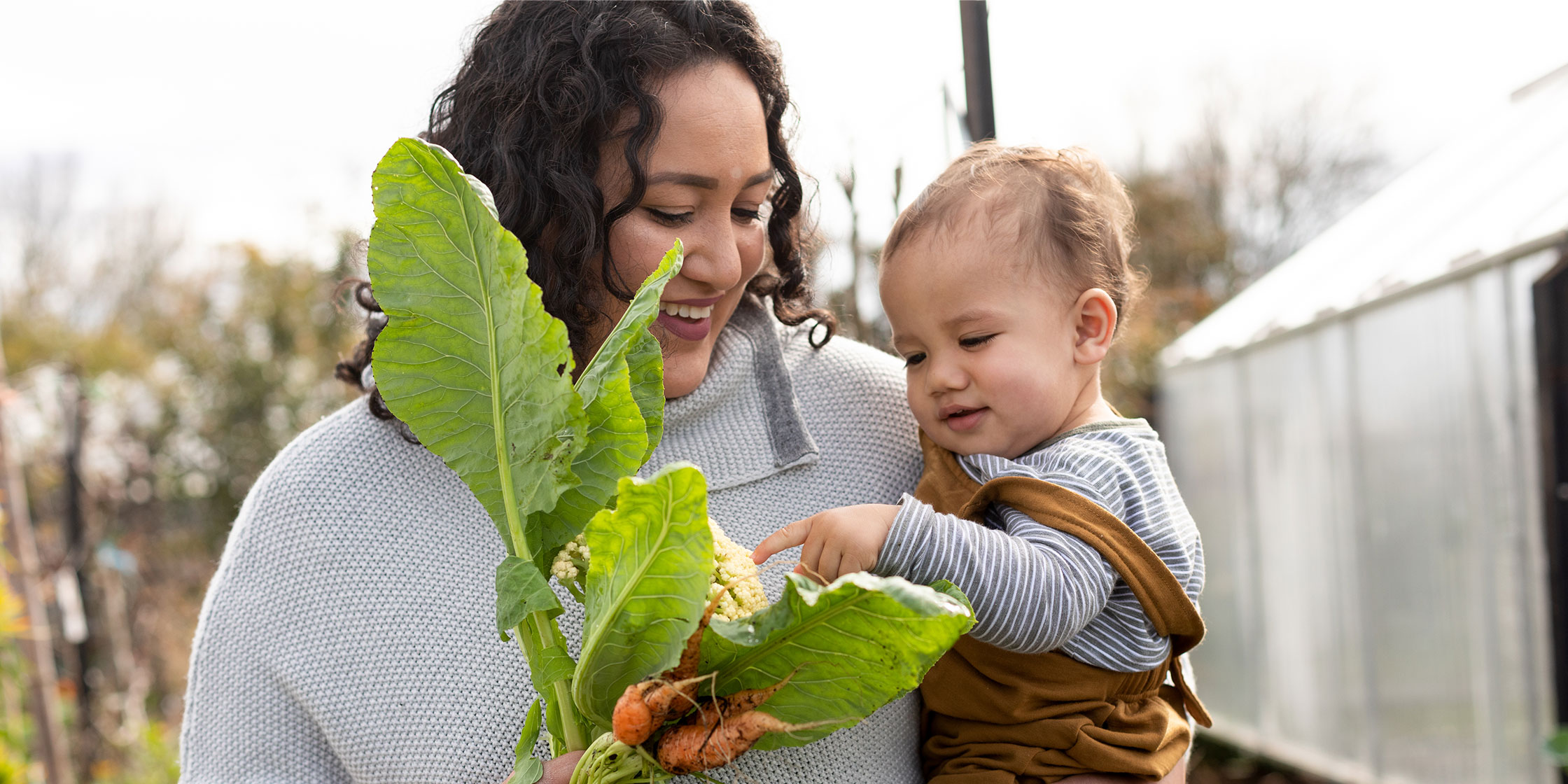 A smiling mother holding her baby while looking at vegetables grown in their garden.