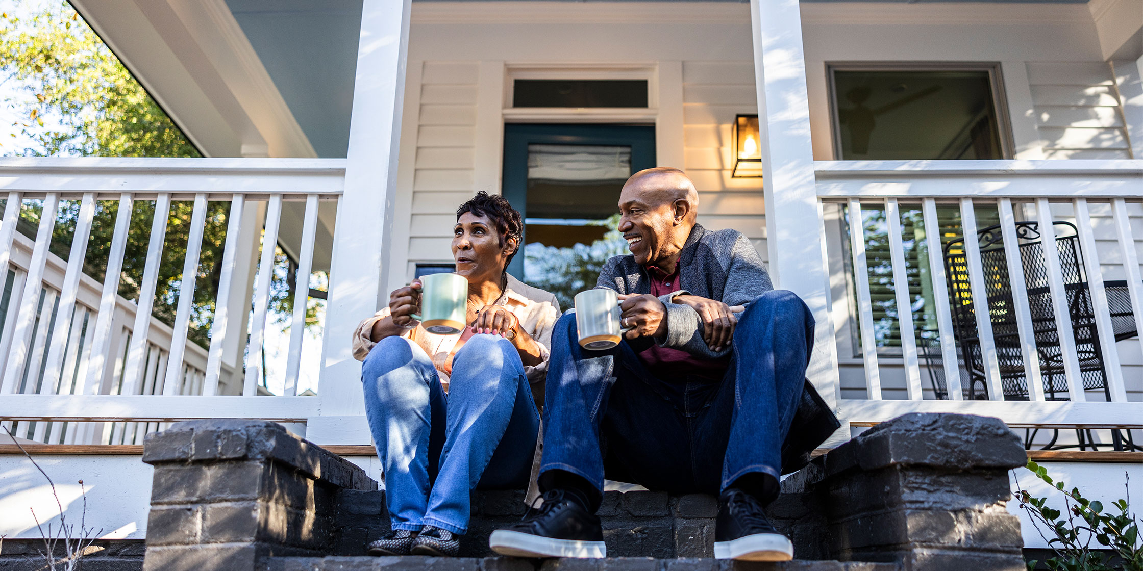 Two people sit on the front stoop of a house with mugs in their hands.