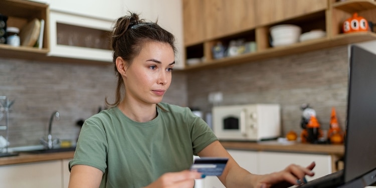 A woman sits at a kitchen table holding her credit card and working on a laptop.