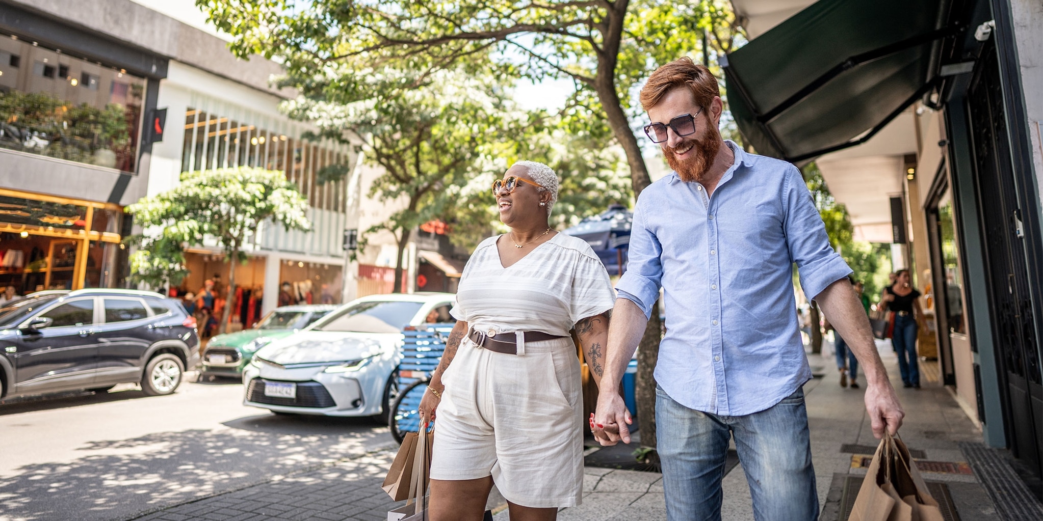 A couple with shopping bags holds hands while walking down a street lined with retail stores.