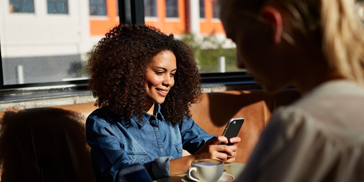A person checks her phone and smiles while sitting at a table with a hot drink.