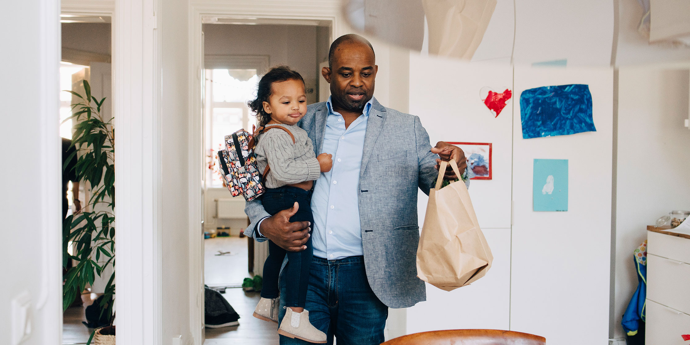 A person is holding a child and carrying a paper bag into another room.