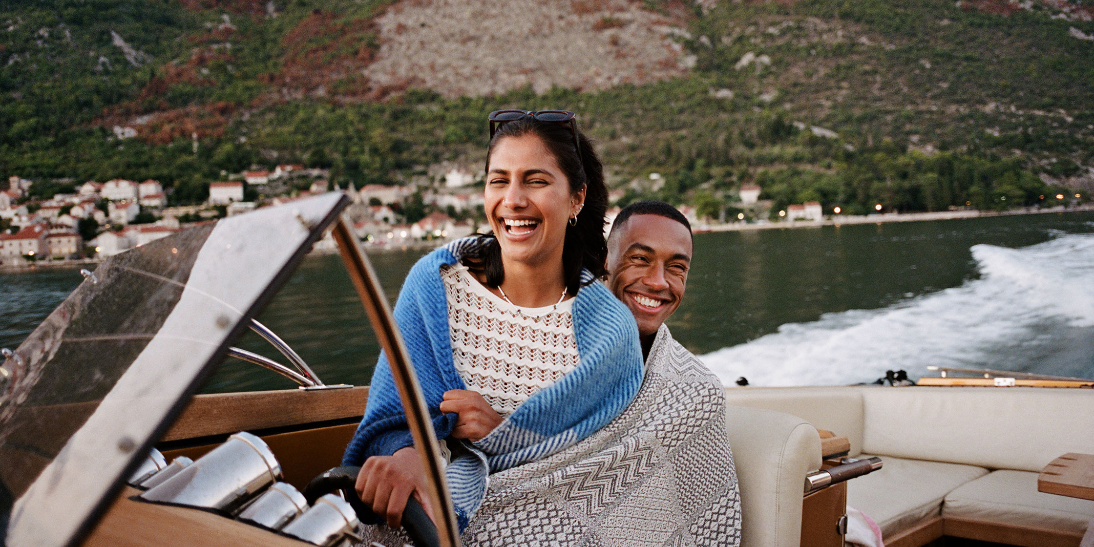 A smiling man and woman wrapped in a blanket steer a wooden speedboat along the Italian coastline.