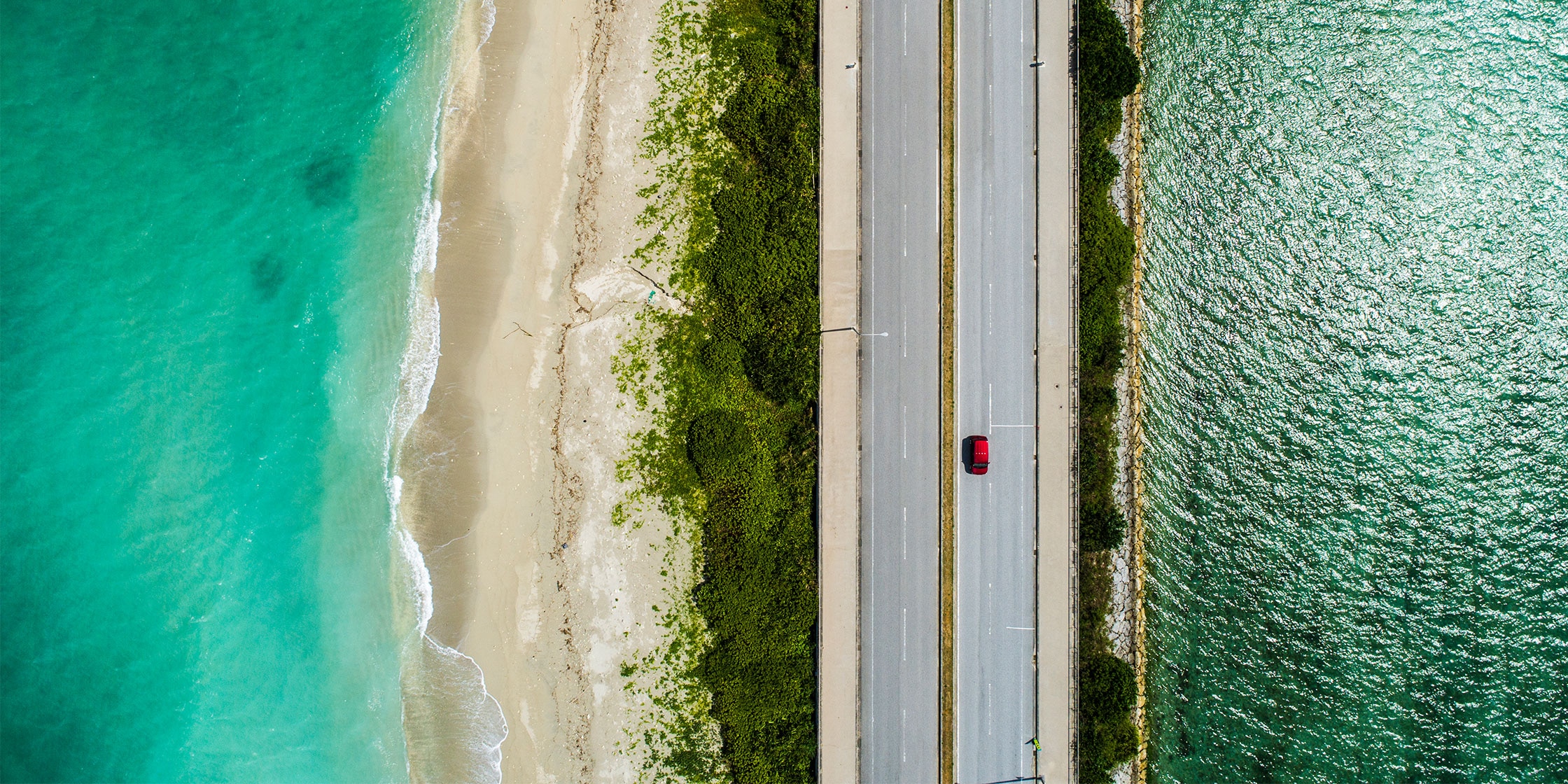 A photo shot from above of a car driving on an open road with water on both sides.