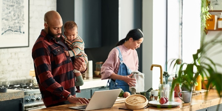 A person standing over a laptop holds a young child in a kitchen while another person dries dishes.