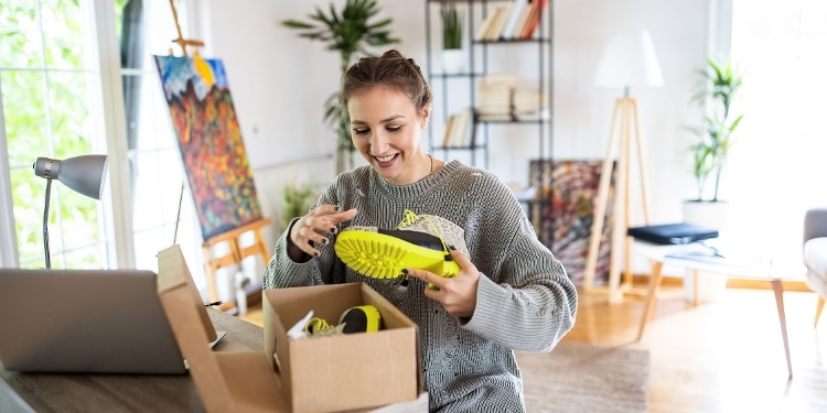 A person in a living room opens a box from Amazon.com with new shoes inside.