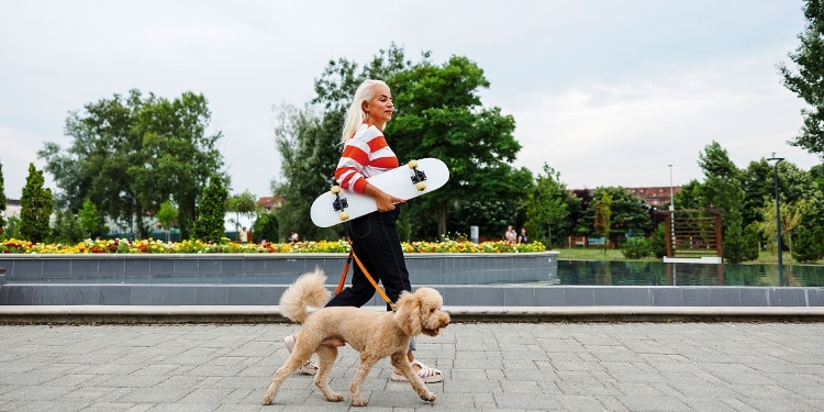 A woman in a striped sweater carrying her skateboard while she walks her dog.