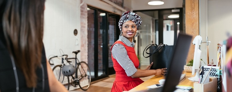 Two women stand beside each other in front of their computers at their shared workspace. One woman looks at the other and smiles.