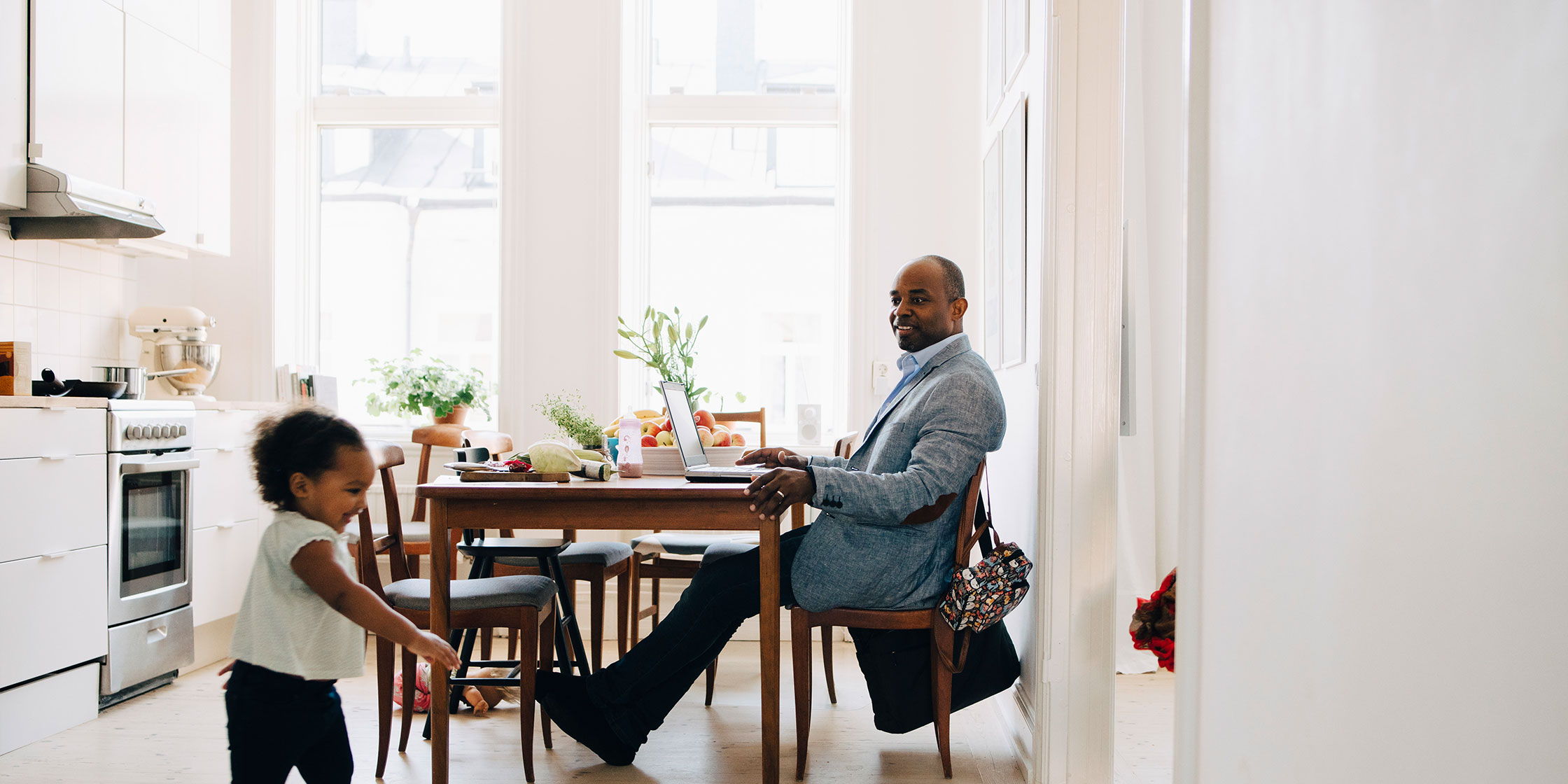 A smiling father sitting at the kitchen table and watching his young daughter run around the kitchen.