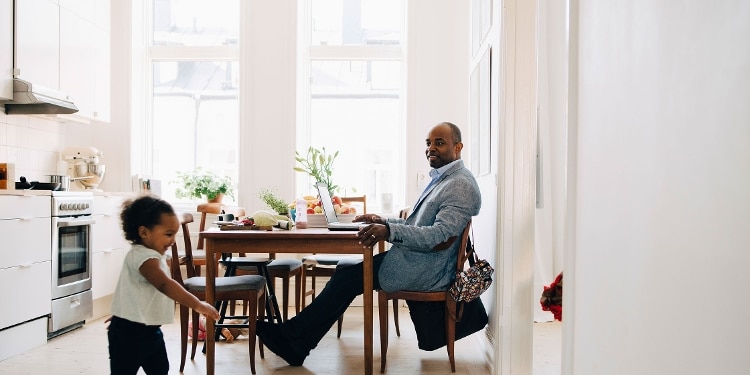 A smiling father sitting at the kitchen table and watching his young daughter run around the kitchen.