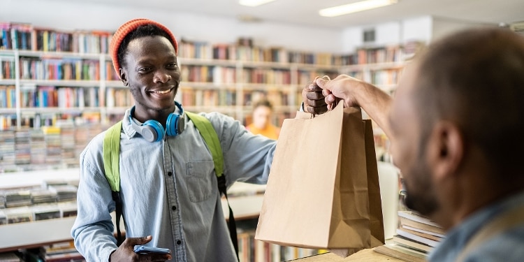 A cashier hands a bag of goods to a young man wearing a hat and headphones.