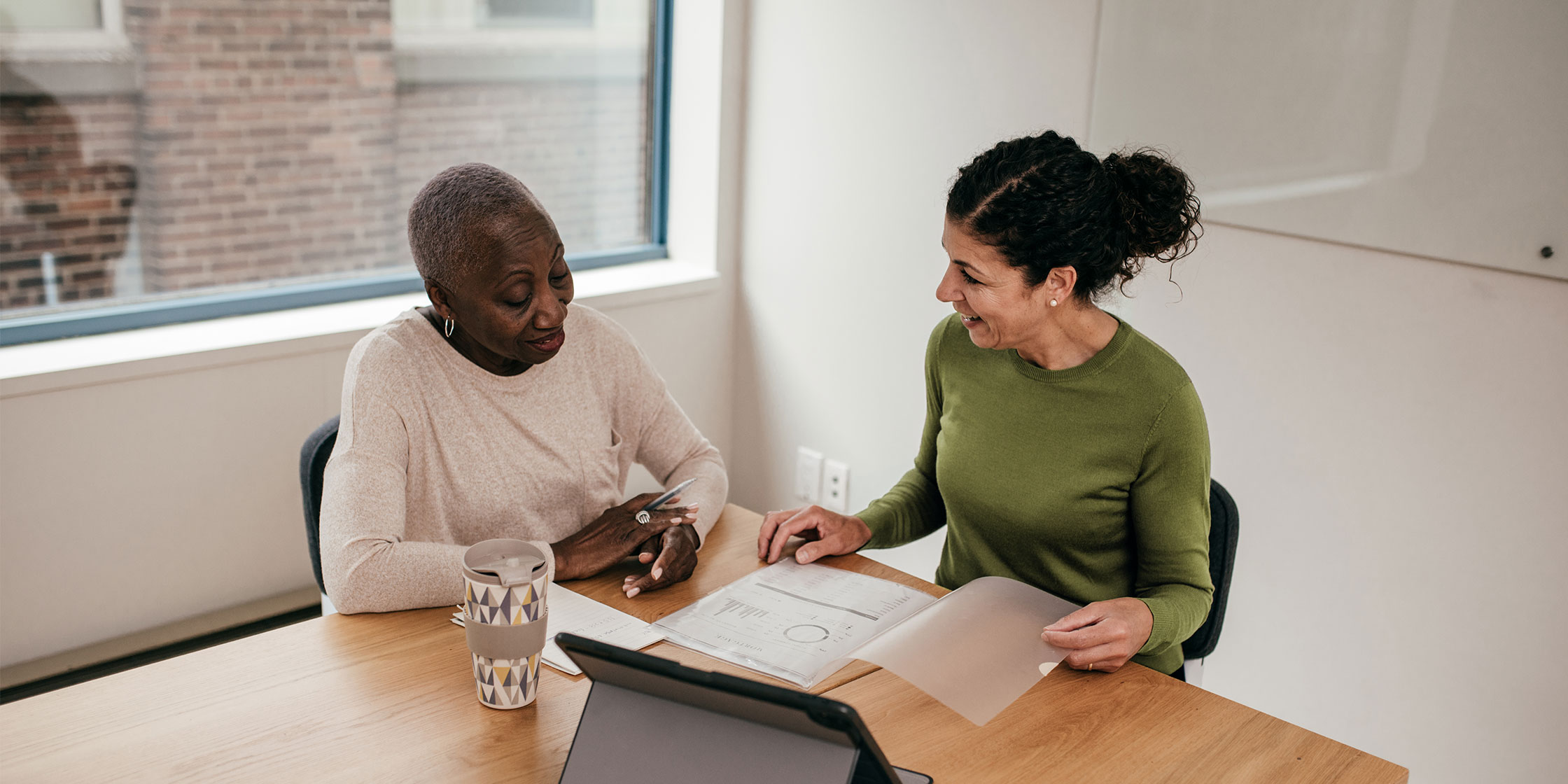 Two people sit at a table discussing paperwork with a tablet nearby.