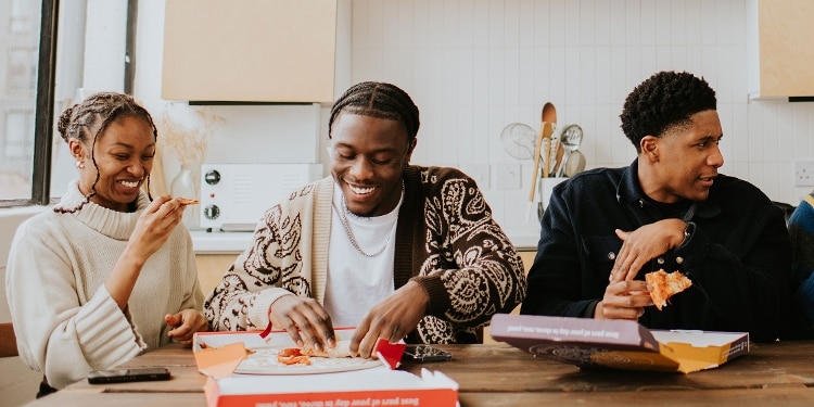 Three students eat pizza and laugh around a kitchen table.
