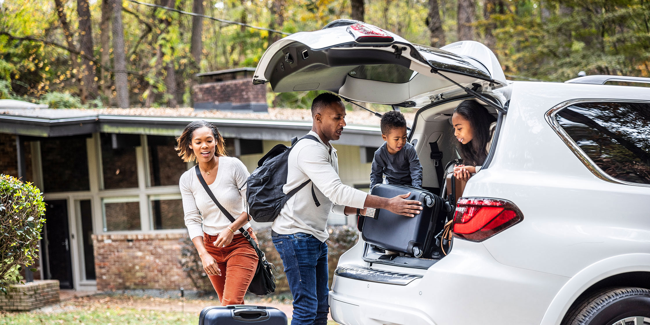 A man and a woman load the trunk of a car with suitcases while two children help from inside the car.