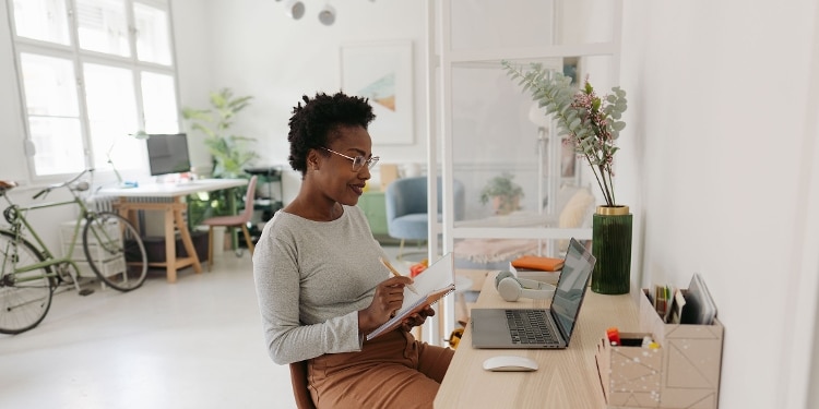 A smiling person in glasses sitting at their desk and looking at their laptop while taking notes.