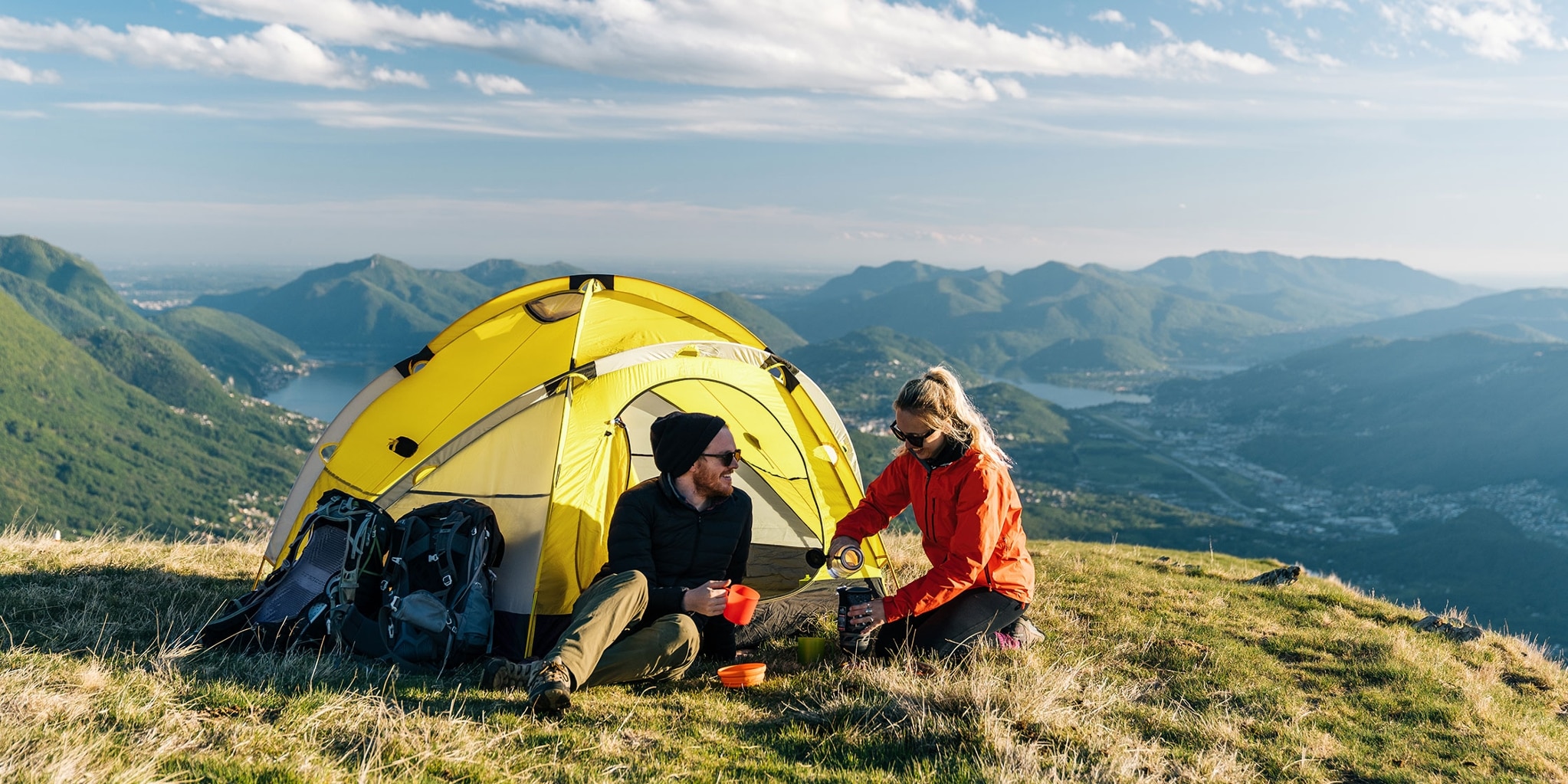 A couple sit outside a tent pitched at the top of a mountain, preparing and enjoying a meal.