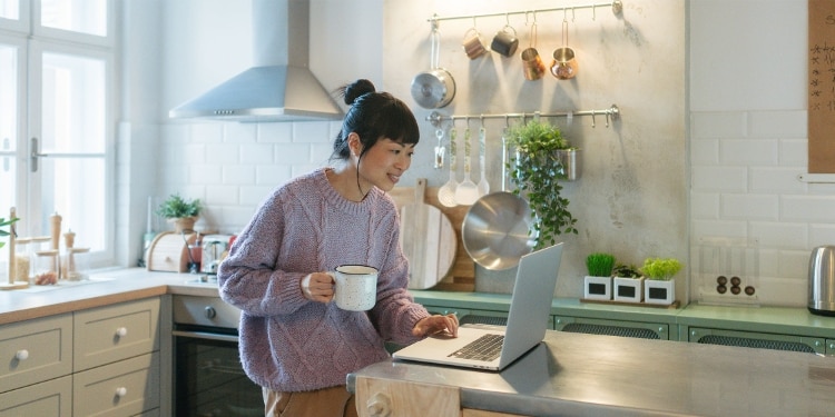A person uses a laptop to search for instant-use credit cards while drinking coffee in a kitchen.
