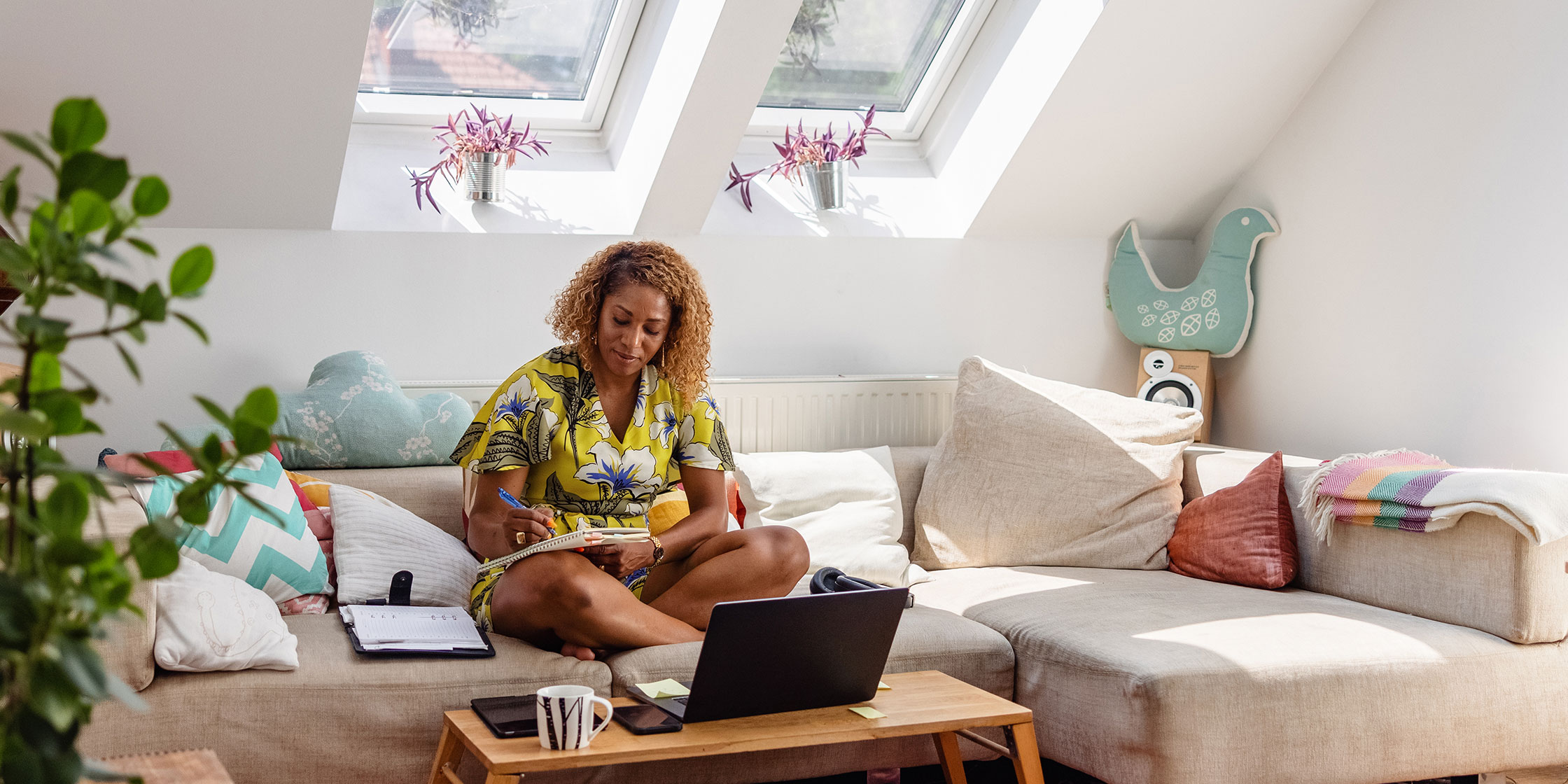 A person at home sits on their couch with an open laptop, making notes in a notebook.