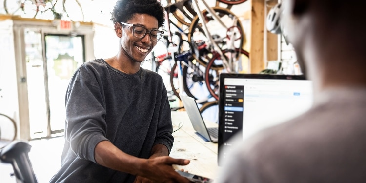 A smiling person wearing glasses looks at their phone while their friend looks at their laptop.