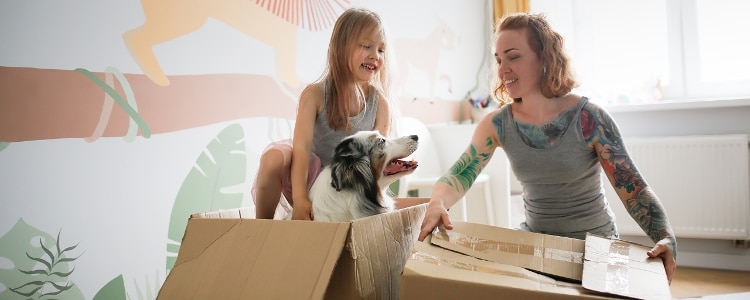 A parent and child unpack boxes with their dog in their new home.