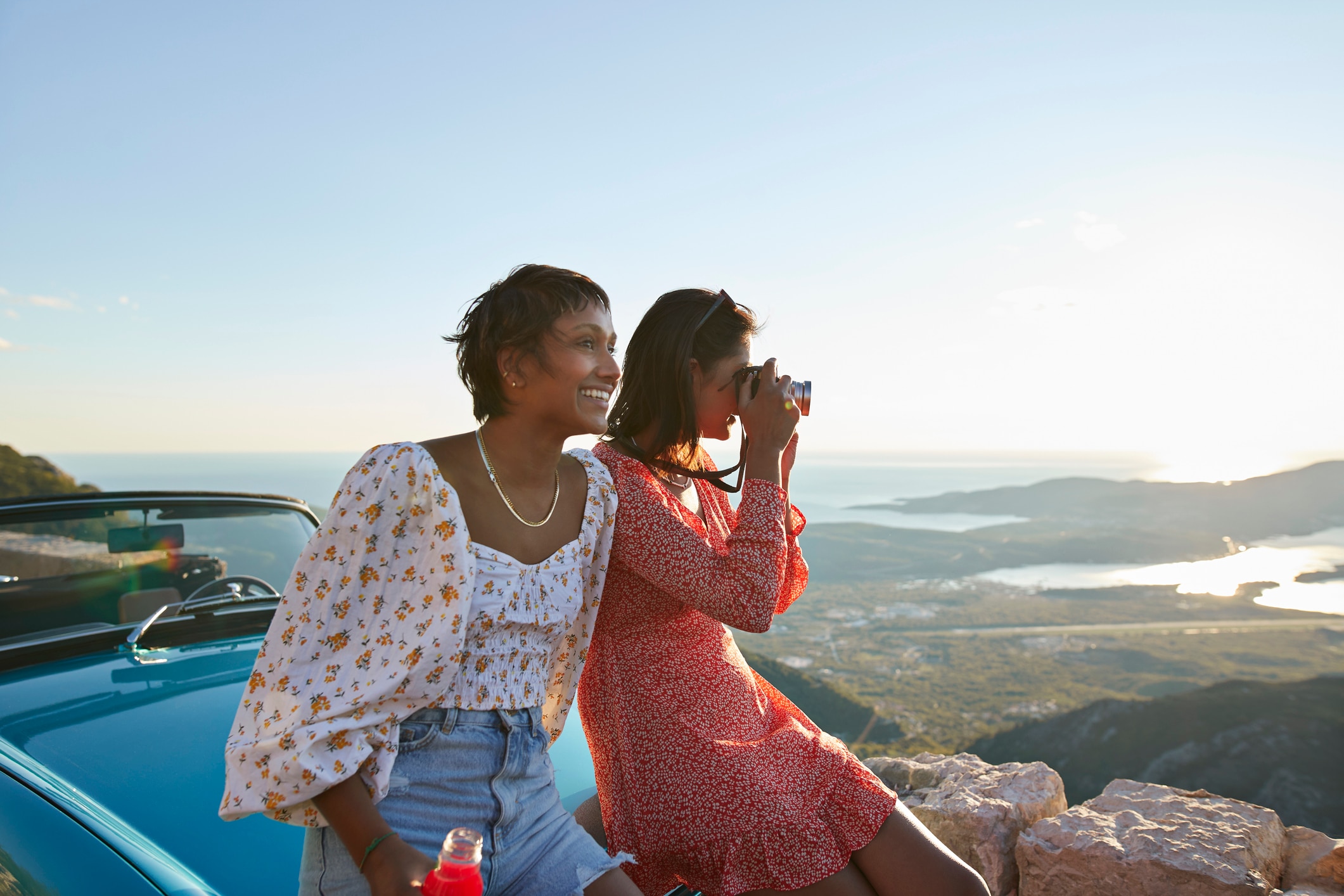 Two women sit on the hood of a car looking out at a rugged coastline.
