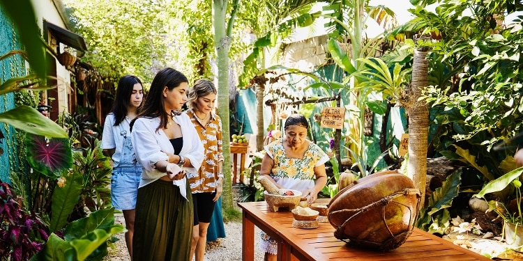 Three travelers watch a traditional Mayan cooking class in a lush garden.