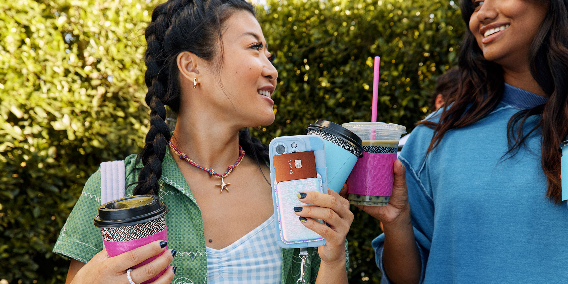 Two friends enjoy their coffee outdoors, one holding a Savor Student card in their smartphone case.