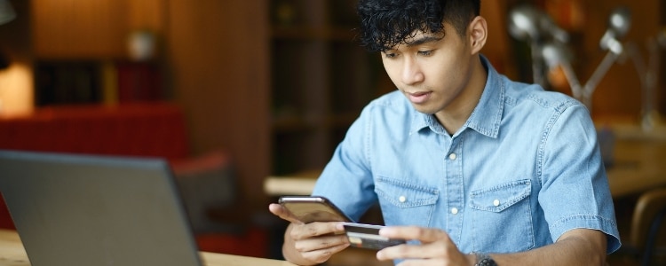 A person sits at their laptop while holding their phone in one hand and a credit card in the other.