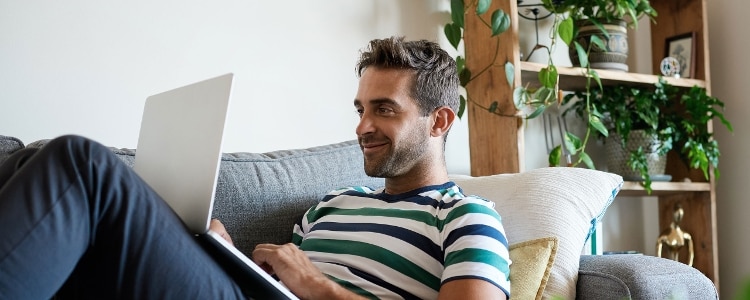 A smiling person looks at their laptop while reclining on a couch.
