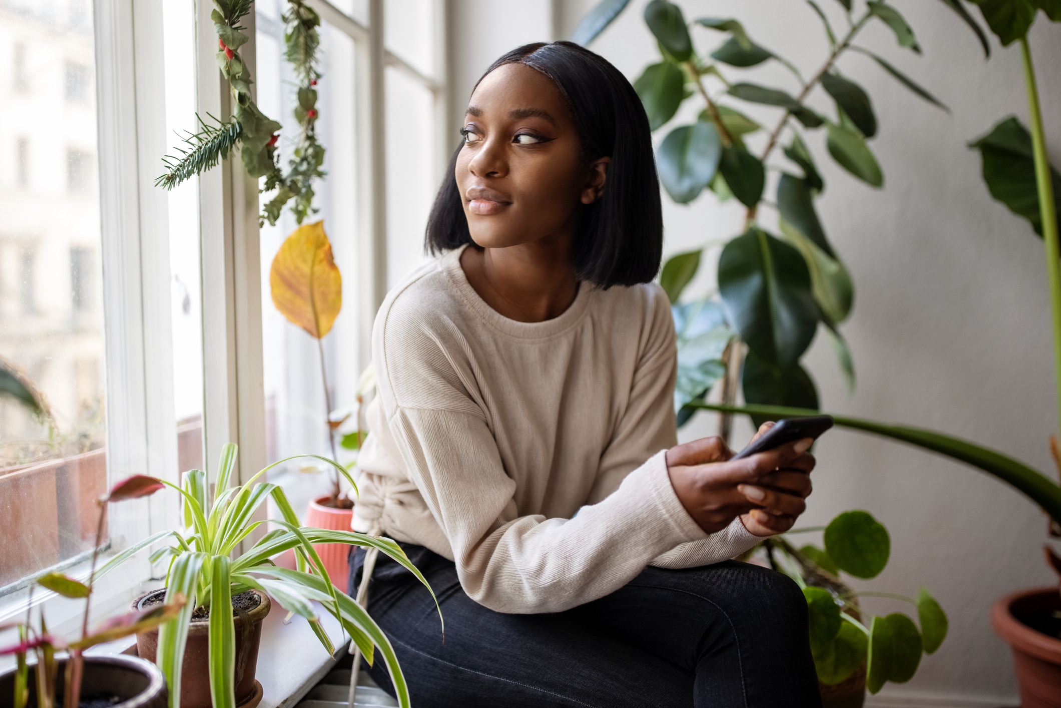 A woman in a plant-filled room looks out the window before she places an online order on her phone.