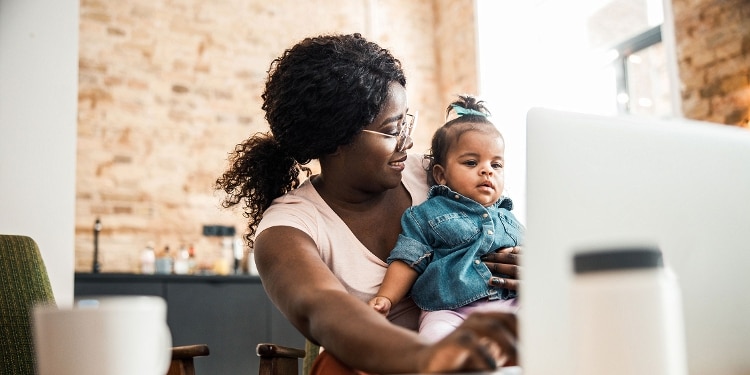 A smiling mother in glasses holding her baby while working on her computer.