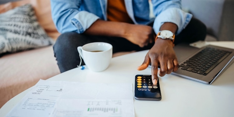 A person sits on a couch with their laptop and a cup of tea, using the calculator app on their phone.