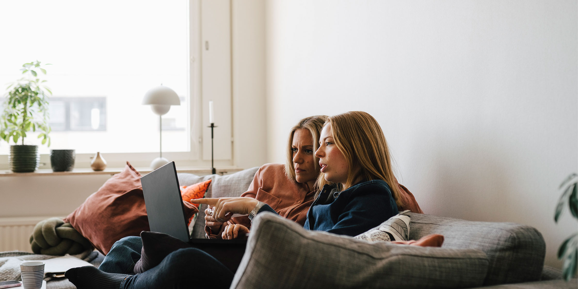 Two women sit on a gray couch, looking at a laptop together while having a conversation.