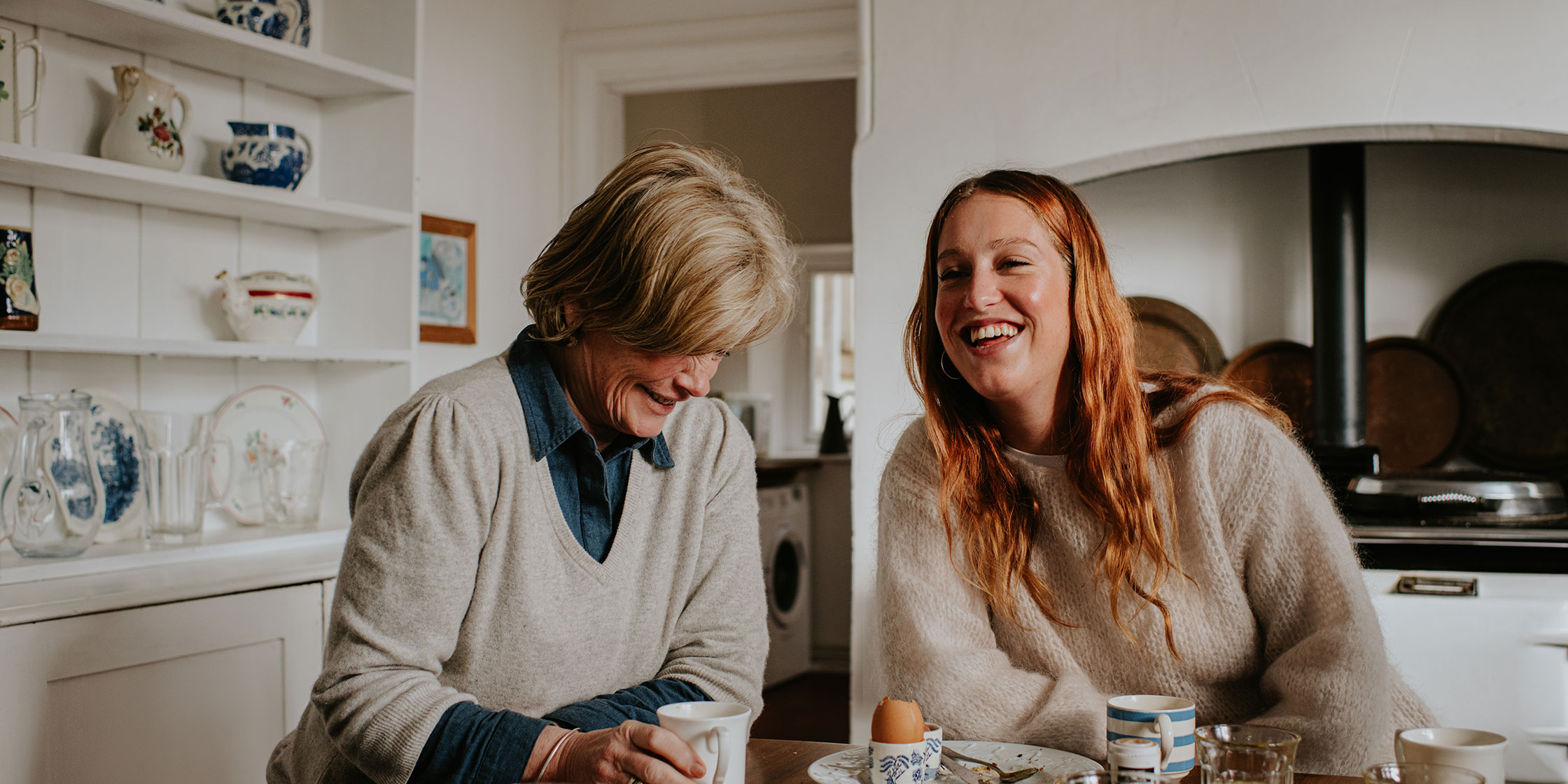 Two women sit at a kitchen table and laugh.