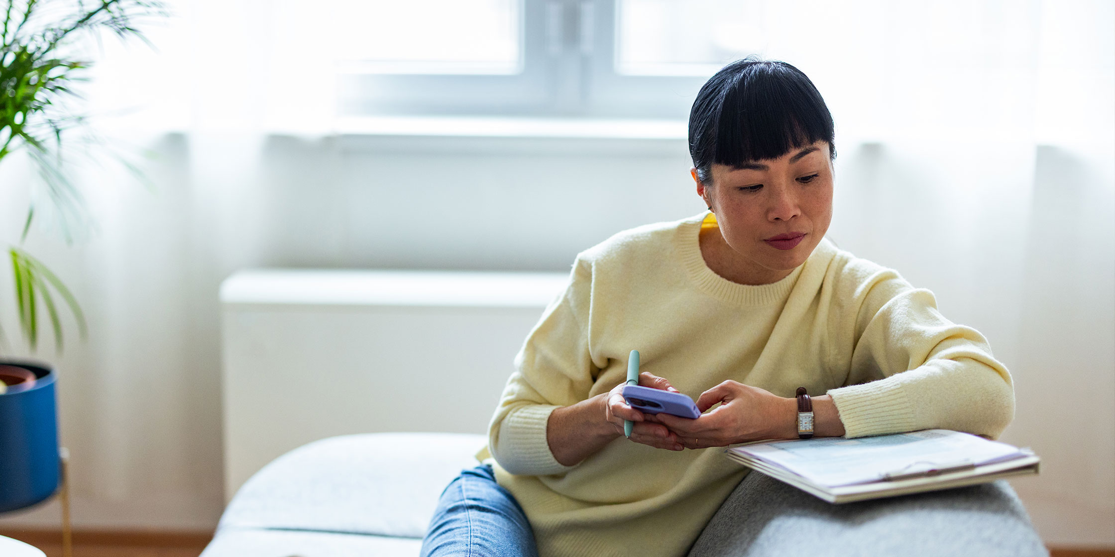 A woman sits in a living room making notes and typing on her phone.