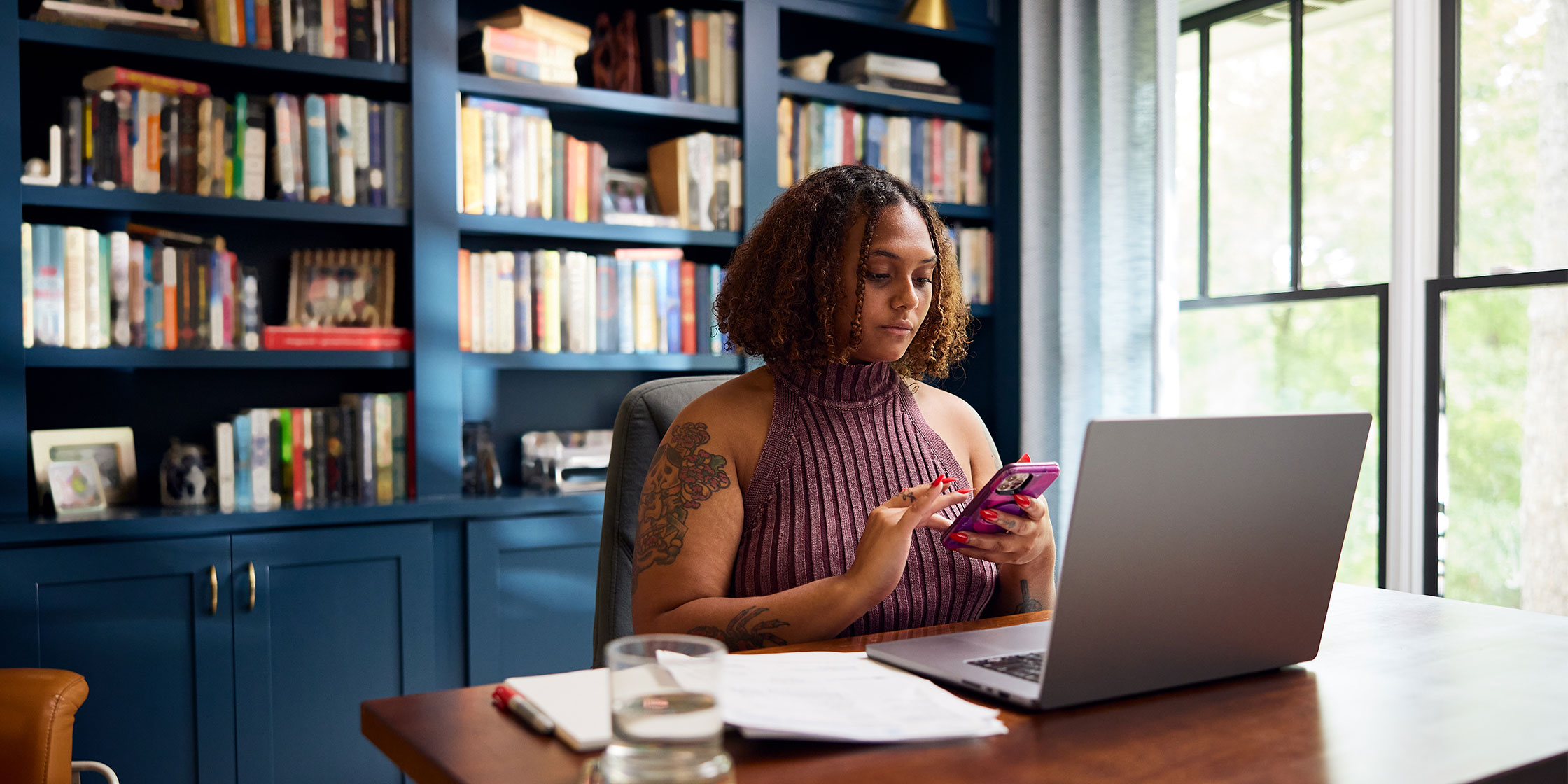 A woman sits at a table in front of a laptop and consults her phone.