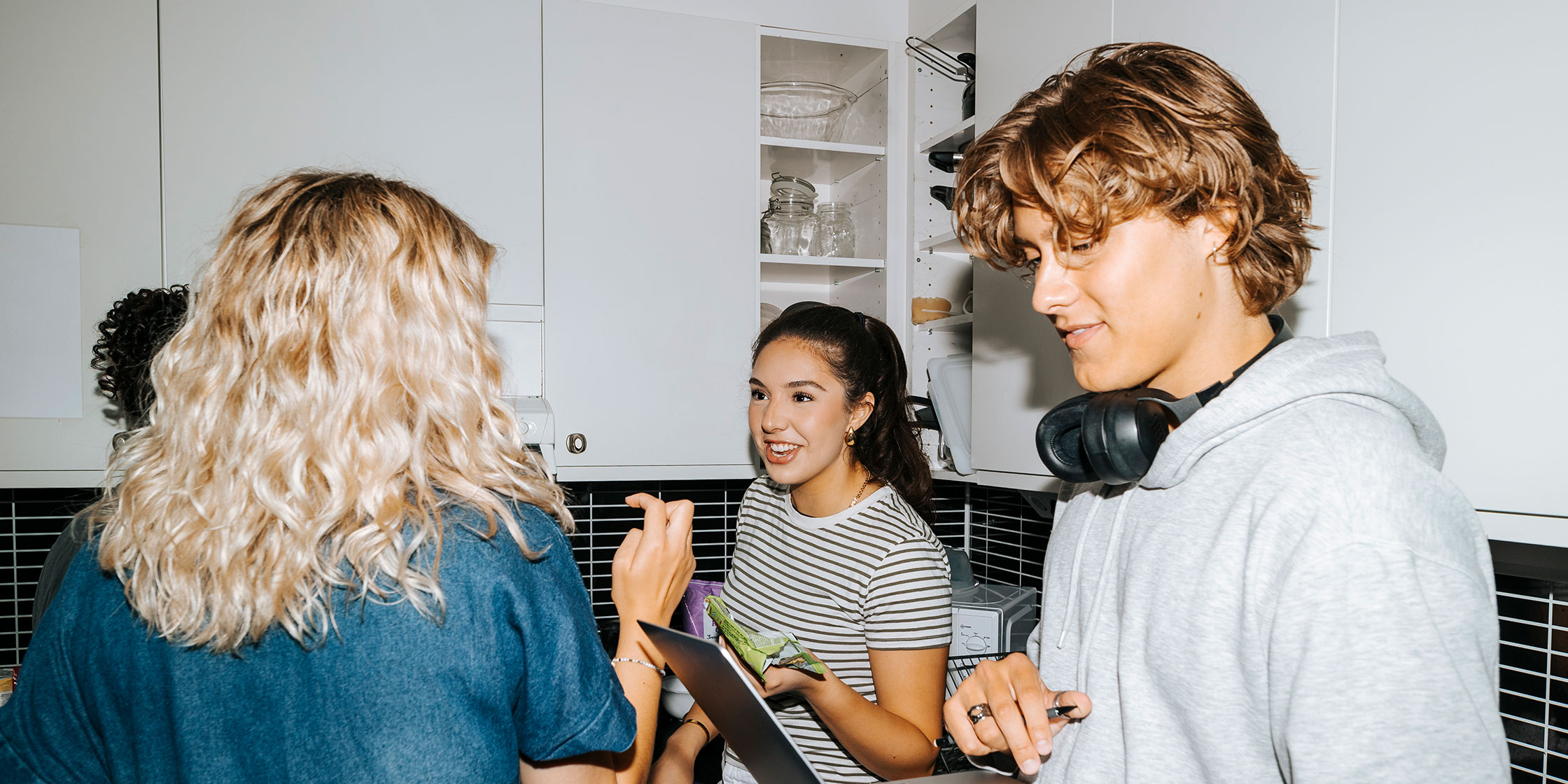 A college student uses a laptop while surrounded by friends in a kitchen.
