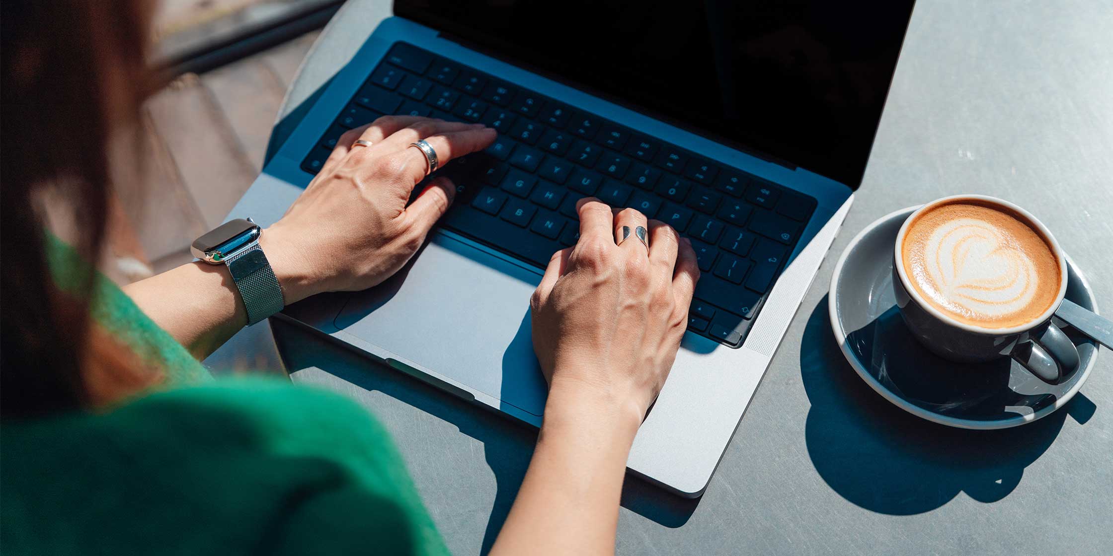 An overhead shot of a person’s hands typing on a laptop. They have a coffee drink beside them on the table.
